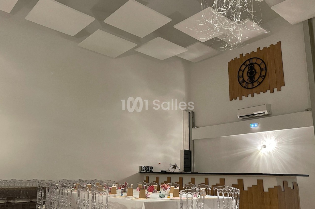 Salle de réception moderne avec tables dressées, chaises transparentes, un bar et une horloge murale décorative.