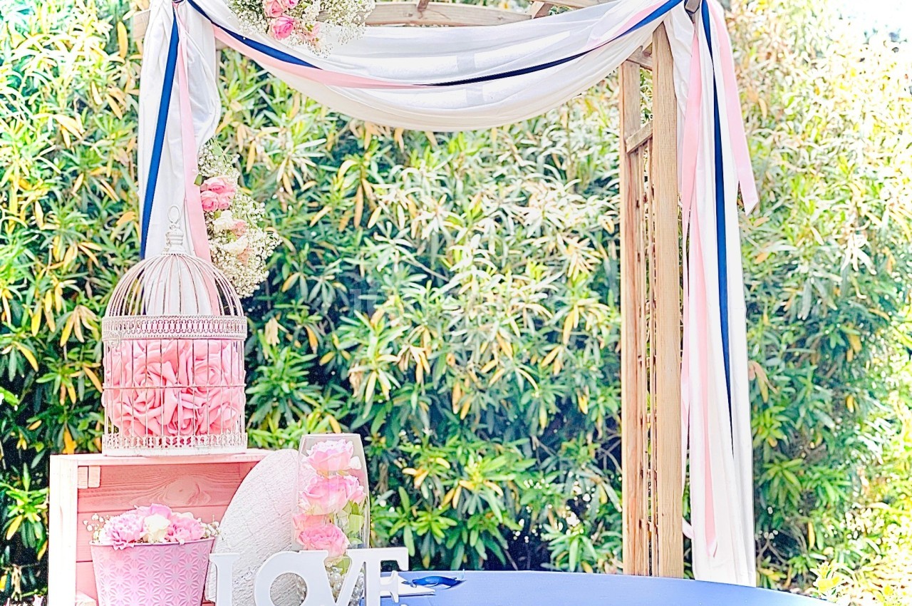 Arche décorée de voilages blancs et roses, avec des fleurs, une cage et des accessoires sur une table bleue.