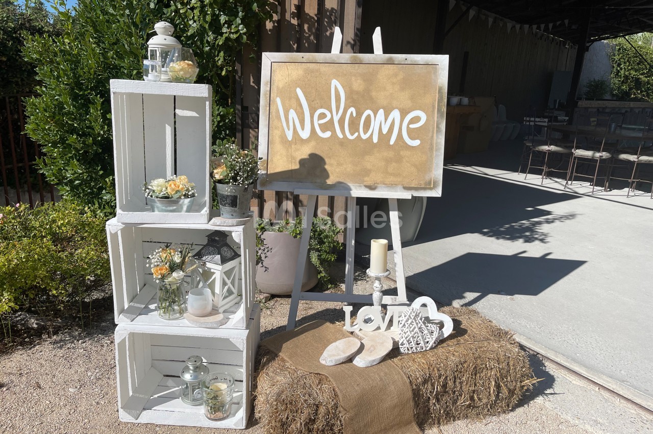 Panneau ’Welcome’ sur chevalet entouré de caisses blanches décorées de bougies, fleurs et objets, dans un cadre extérieur…