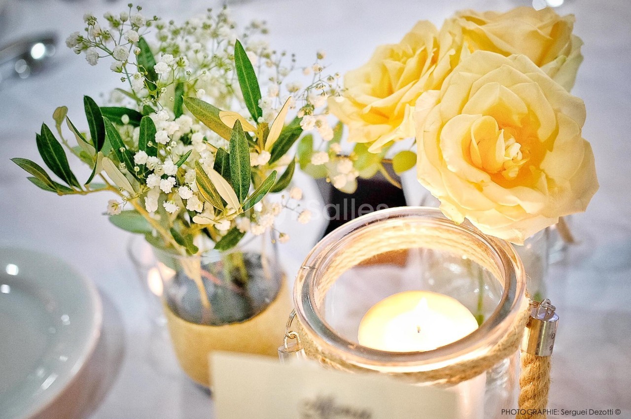 Bouquet de roses jaunes et gypsophiles dans des vases, avec une bougie allumée sur une table blanche.