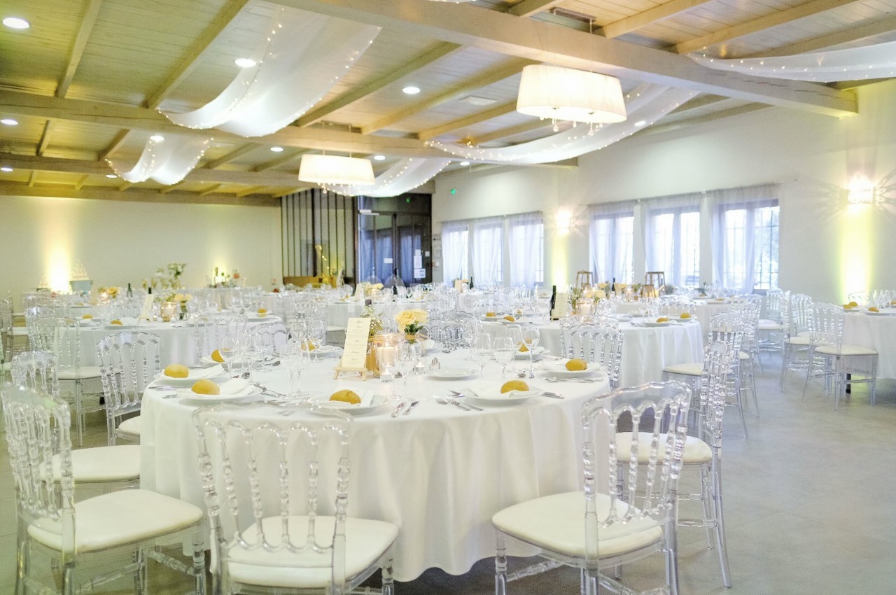 Salle de réception lumineuse avec tables rondes dressées, chaises transparentes et décorations élégantes.