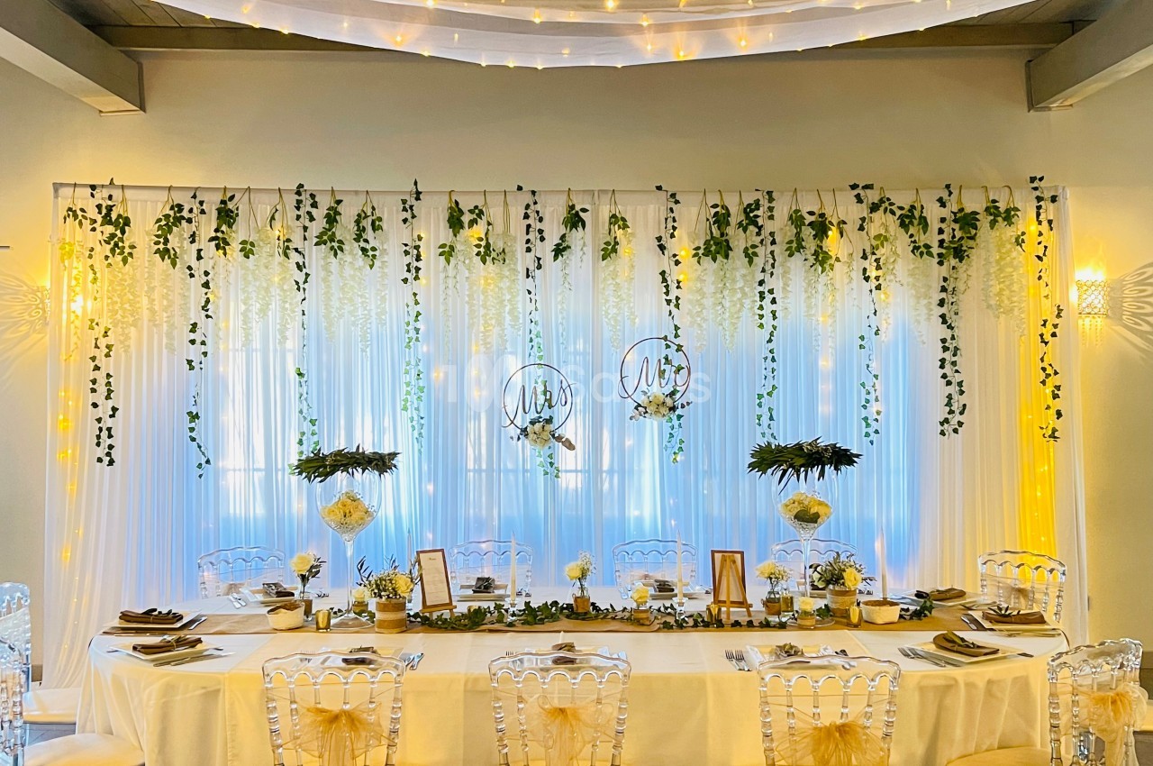Table de réception décorée avec des chaises transparentes, guirlandes lumineuses, feuillages suspendus et fleurs blanches.
