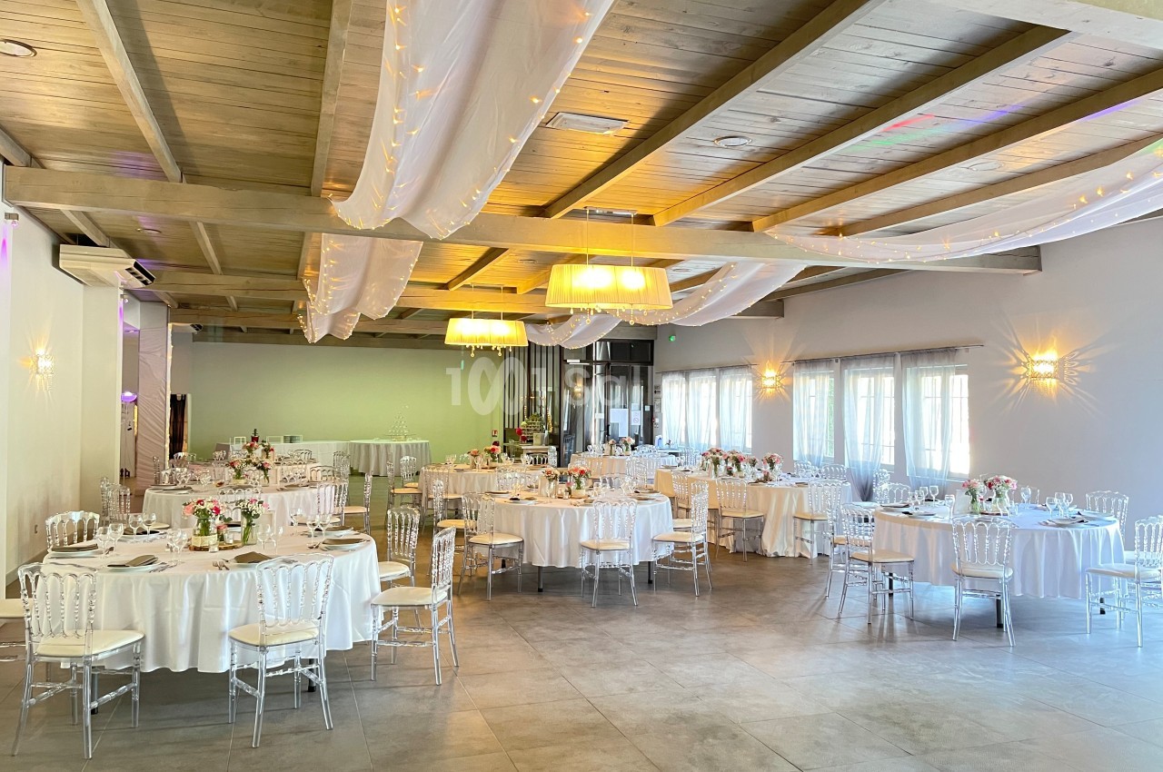 Salle de réception lumineuse avec tables rondes décorées de nappes blanches, chaises transparentes et arrangements floraux.