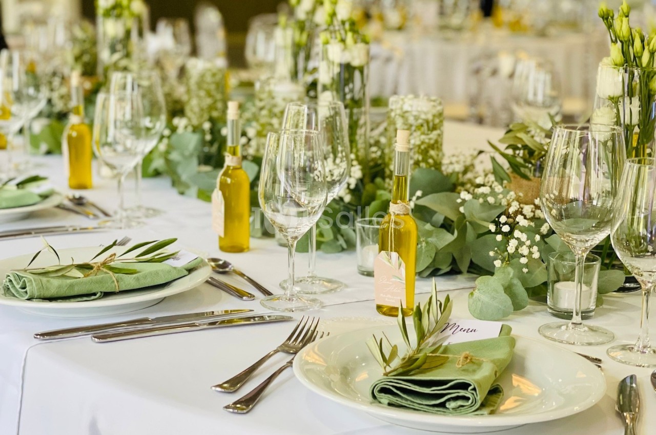 Table élégamment dressée avec nappes blanches, couverts, serviettes vertes, bouteilles d'huile et décorations florales.