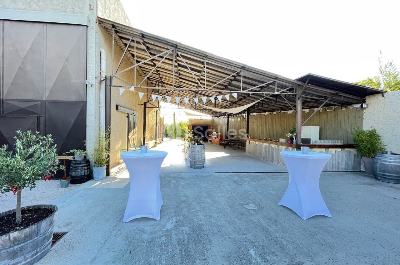 Espace extérieur avec tables hautes drapées de blanc, décoré pour un événement sous une structure métallique ouverte.