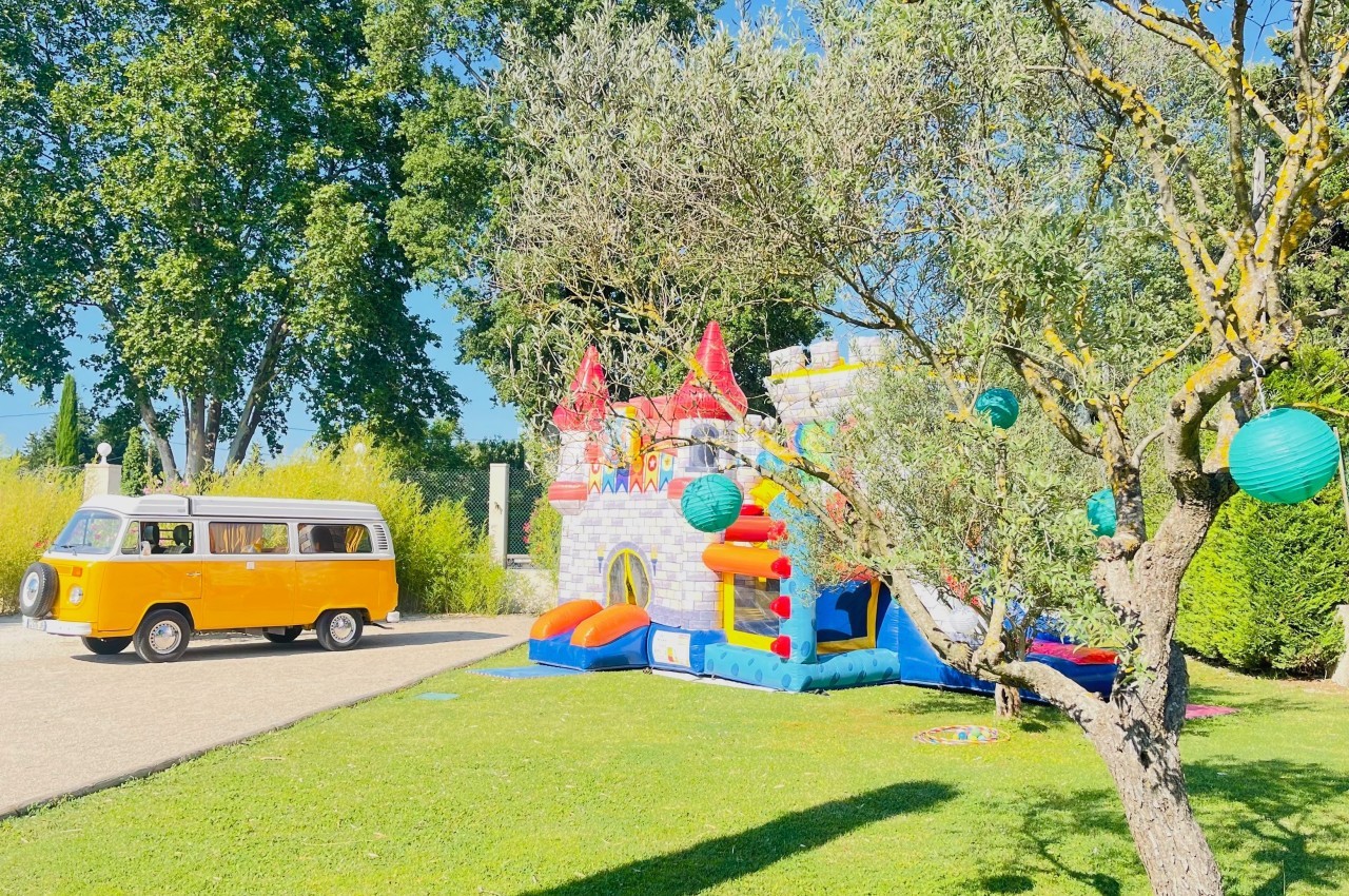 Un combi jaune garé près d'un château gonflable coloré dans un jardin ensoleillé avec des arbres et des lanternes suspendues.