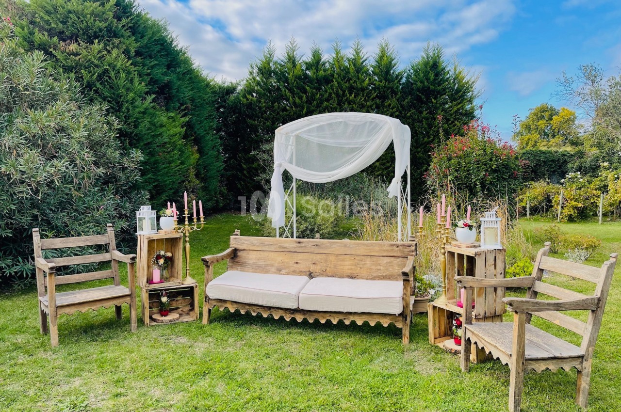 Mobilier en bois avec coussins disposé dans un jardin, entouré de verdure et décoré de bougies et lanternes.