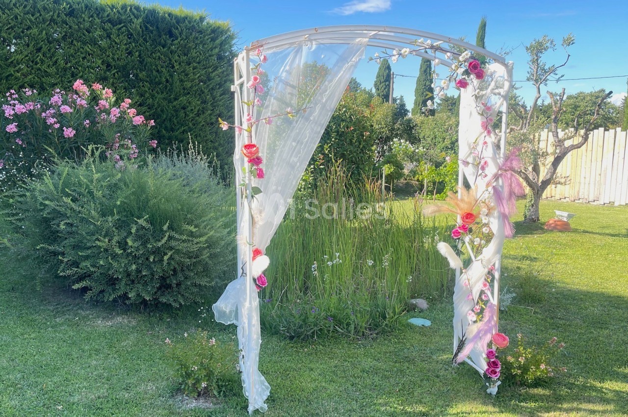 Arche de mariage décorée de fleurs et voilage blanc, installée dans un jardin verdoyant par une journée ensoleillée.
