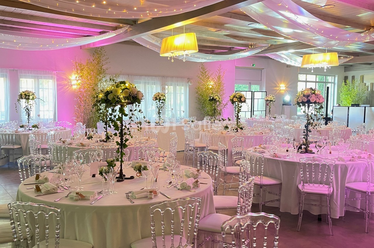 Salle de réception décorée avec des tables rondes, chaises transparentes, fleurs et éclairage rose et blanc.