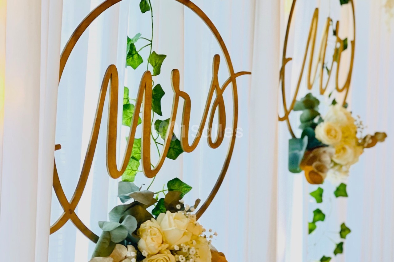 Anneaux décoratifs dorés avec l'inscription ’Mrs’, ornés de fleurs et de feuillage, suspendus devant un rideau blanc.