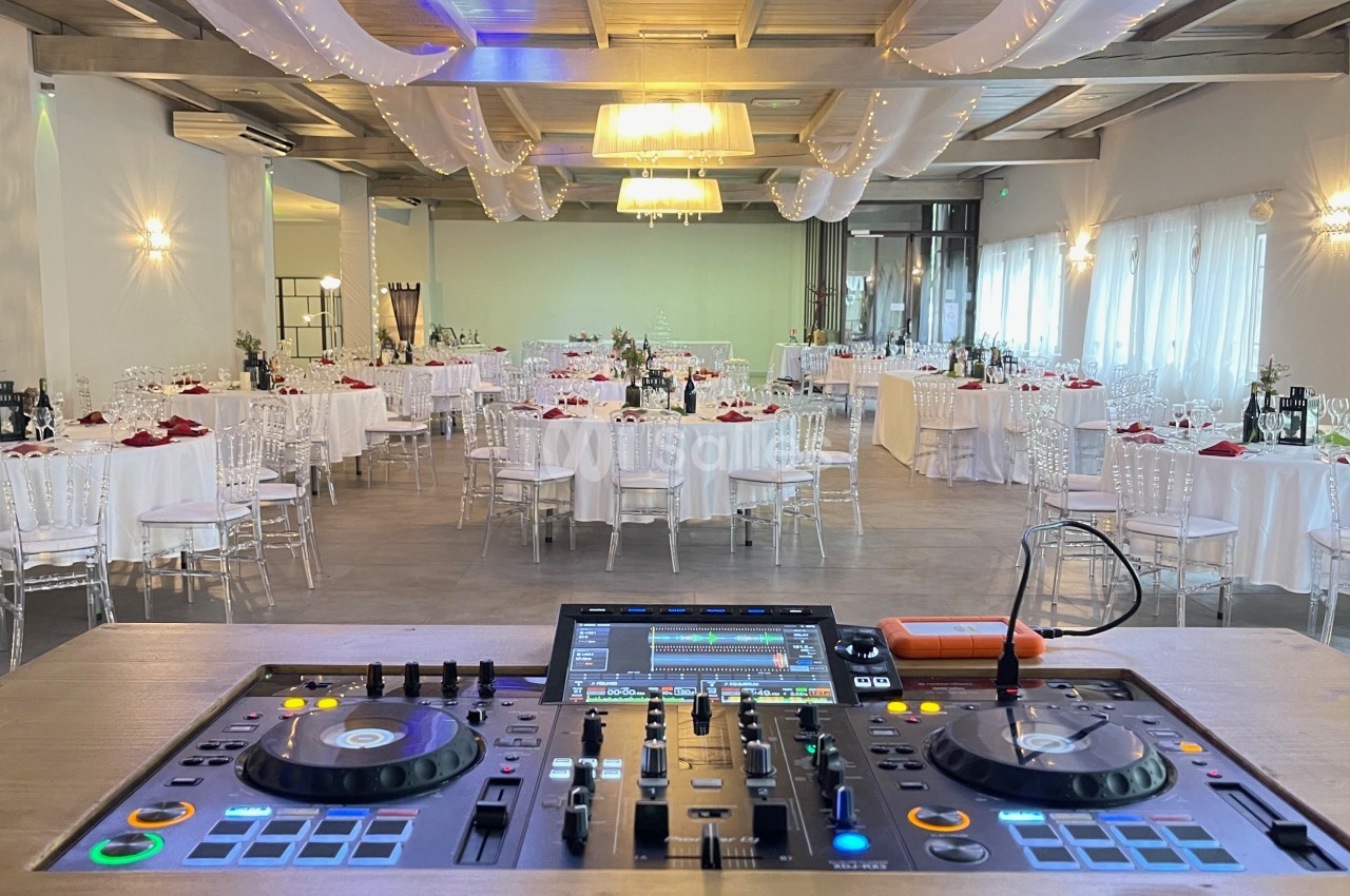Salle de réception décorée avec des tables dressées, vue depuis une table de mixage de DJ au premier plan.