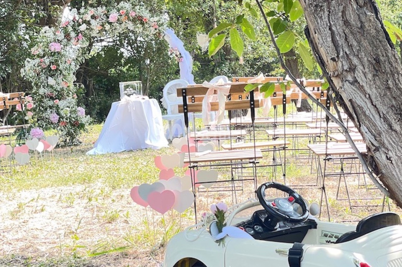 Cérémonie en extérieur avec arche florale, chaises alignées et petite voiture blanche décorée au premier plan.