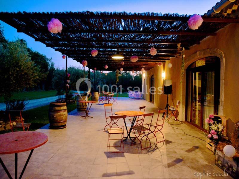 Terrasse extérieure avec tables et chaises en métal, décorée de lanternes et guirlandes sous une pergola au crépuscule.