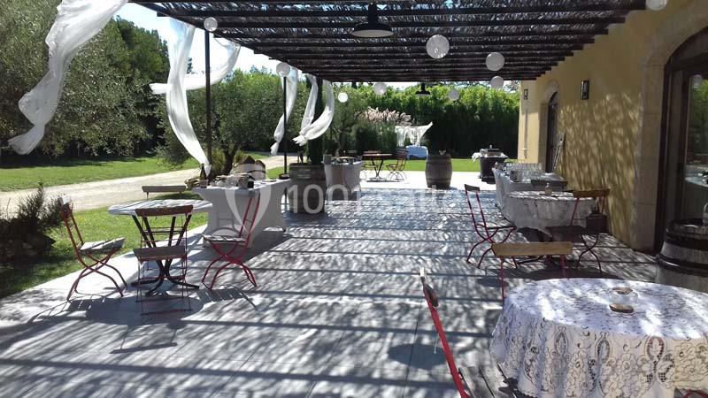 Terrasse ombragée avec tables et chaises, décorée de voilages blancs, entourée de verdure et d'arbres.