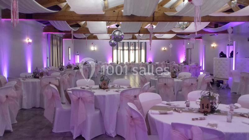 Salle de réception décorée avec des tables rondes, nappes blanches, chaises ornées de nœuds roses et éclairage violet.