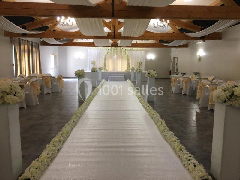 Salle de réception décorée pour un mariage, avec allée centrale bordée de fleurs blanches et chaises ornées de rubans.