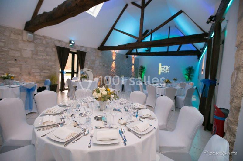 Salle de réception avec murs en pierre, poutres apparentes, tables rondes dressées et éclairage tamisé bleu.