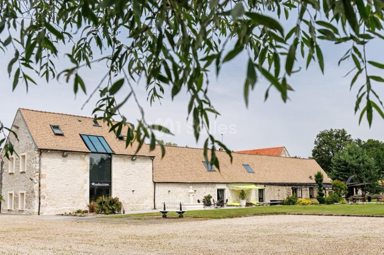 Bâtiment en pierre avec toit en tuiles, entouré d'un jardin, vu à travers des branches d'arbres.