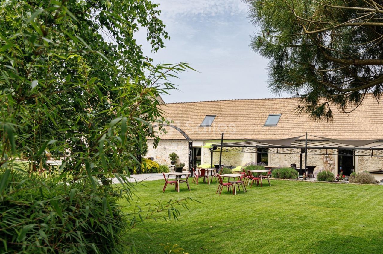 Jardin verdoyant avec pelouse, tables et chaises en bois, entouré de végétation et d'un bâtiment en pierre.