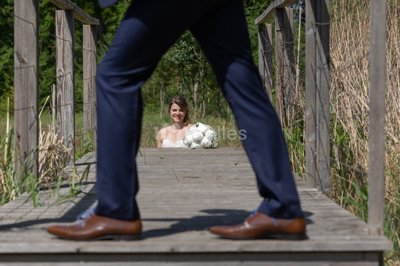 Une mariée souriante tenant un bouquet est visible à travers les jambes d'un homme en costume sur un pont en bois.