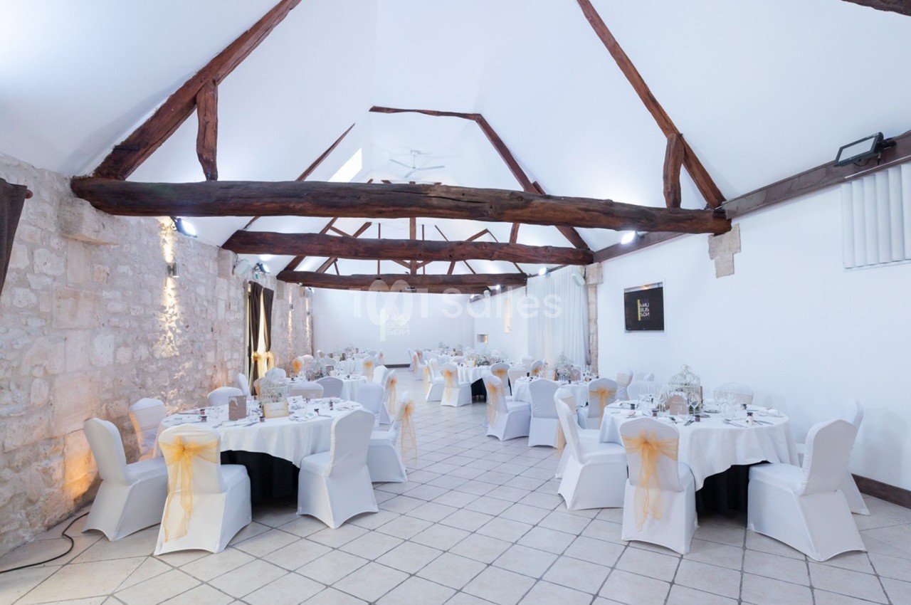 Salle de réception lumineuse avec poutres apparentes, tables rondes dressées et chaises blanches décorées de nœuds.