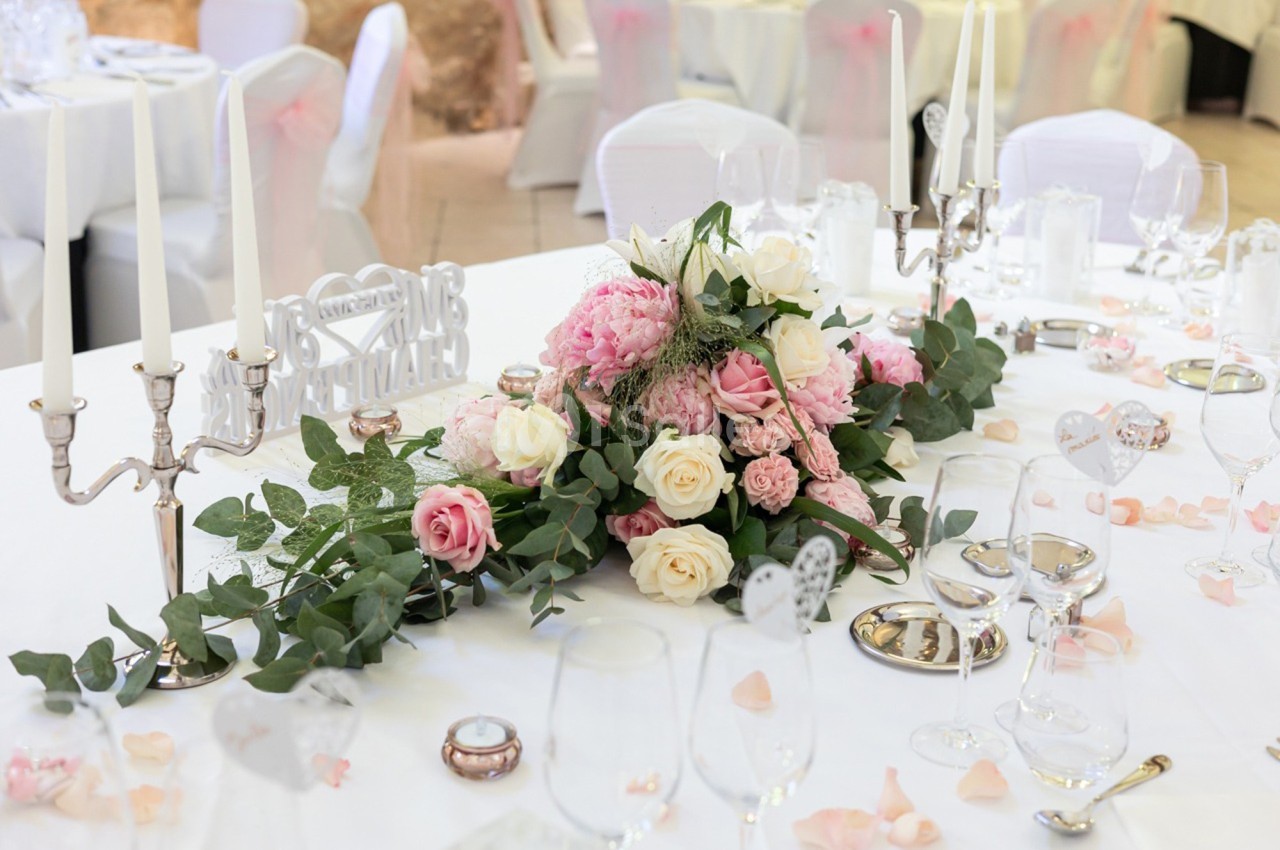 Centre de table floral avec roses et pivoines roses et blanches, entouré de bougies et vaisselle sur une nappe blanche.