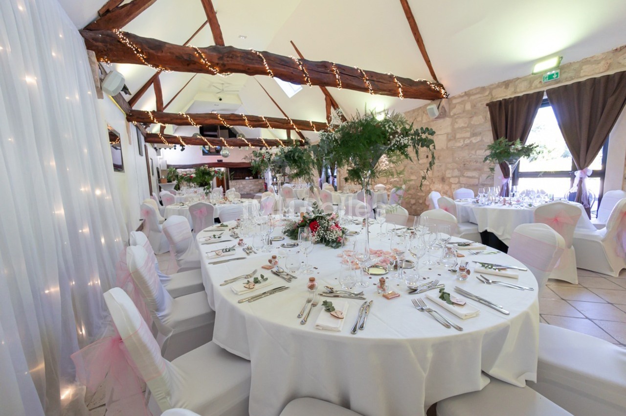 Salle de réception décorée avec des tables rondes dressées, chaises blanches et guirlandes lumineuses au plafond.