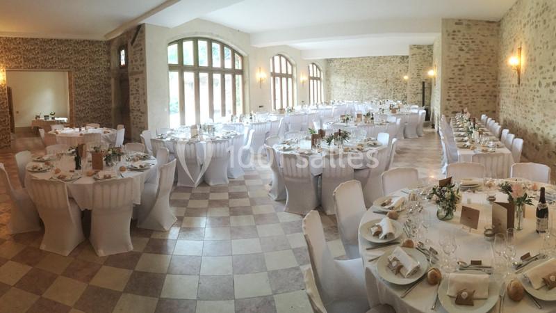Salle de réception lumineuse avec des tables rondes dressées pour un événement, entourées de chaises blanches.