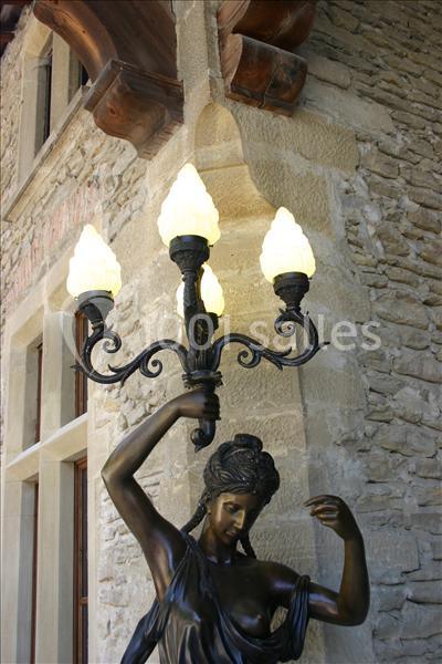 Statue en bronze représentant une femme tenant un lampadaire à quatre globes lumineux devant un mur en pierre.