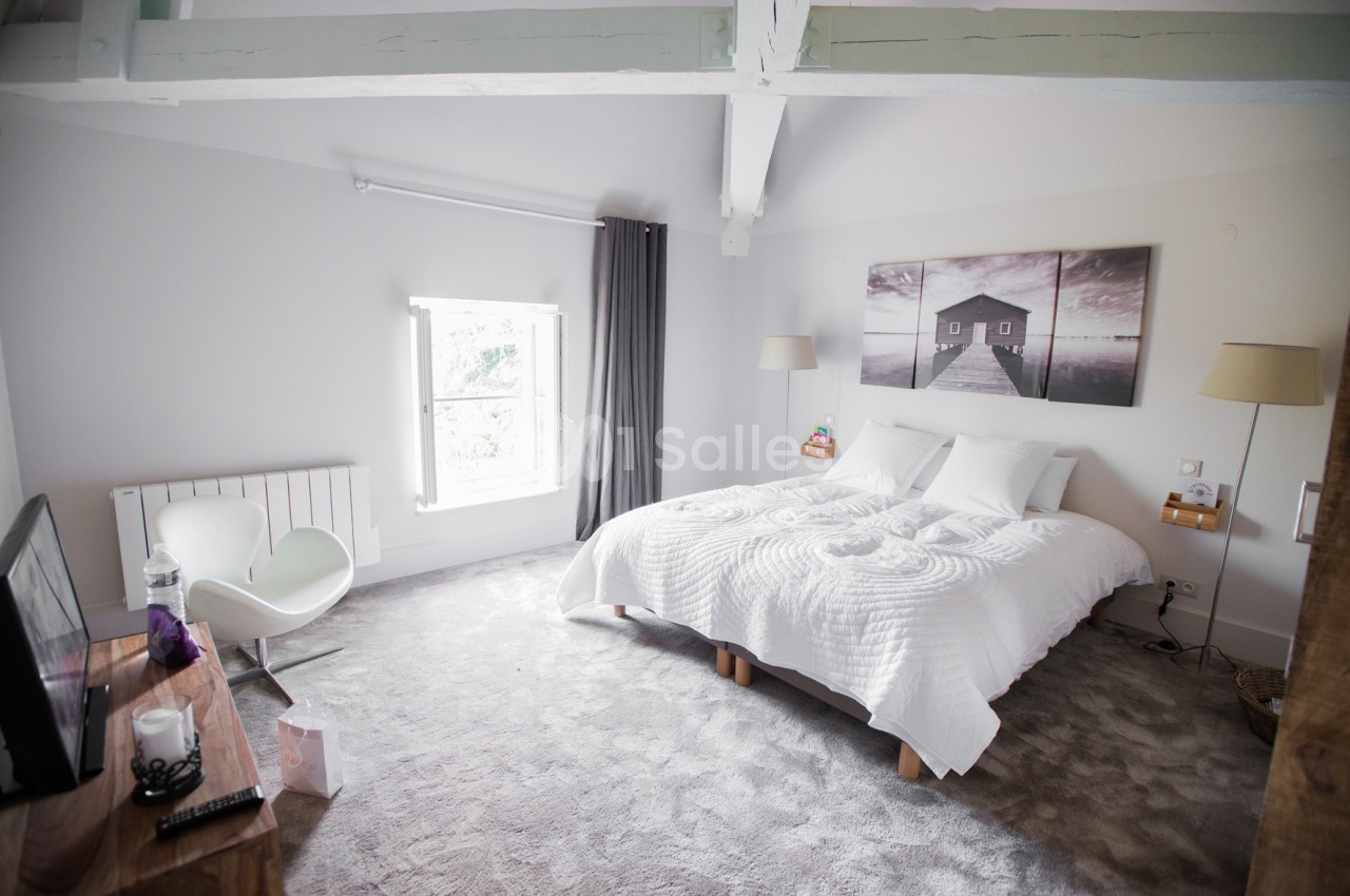 Chambre lumineuse avec lit double, murs blancs, plafond avec poutres apparentes et grande fenêtre avec rideaux.