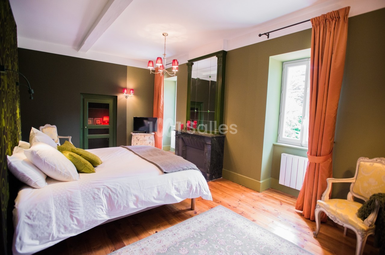 Chambre lumineuse avec lit double, murs verts, cheminée, rideaux orange et parquet en bois.
