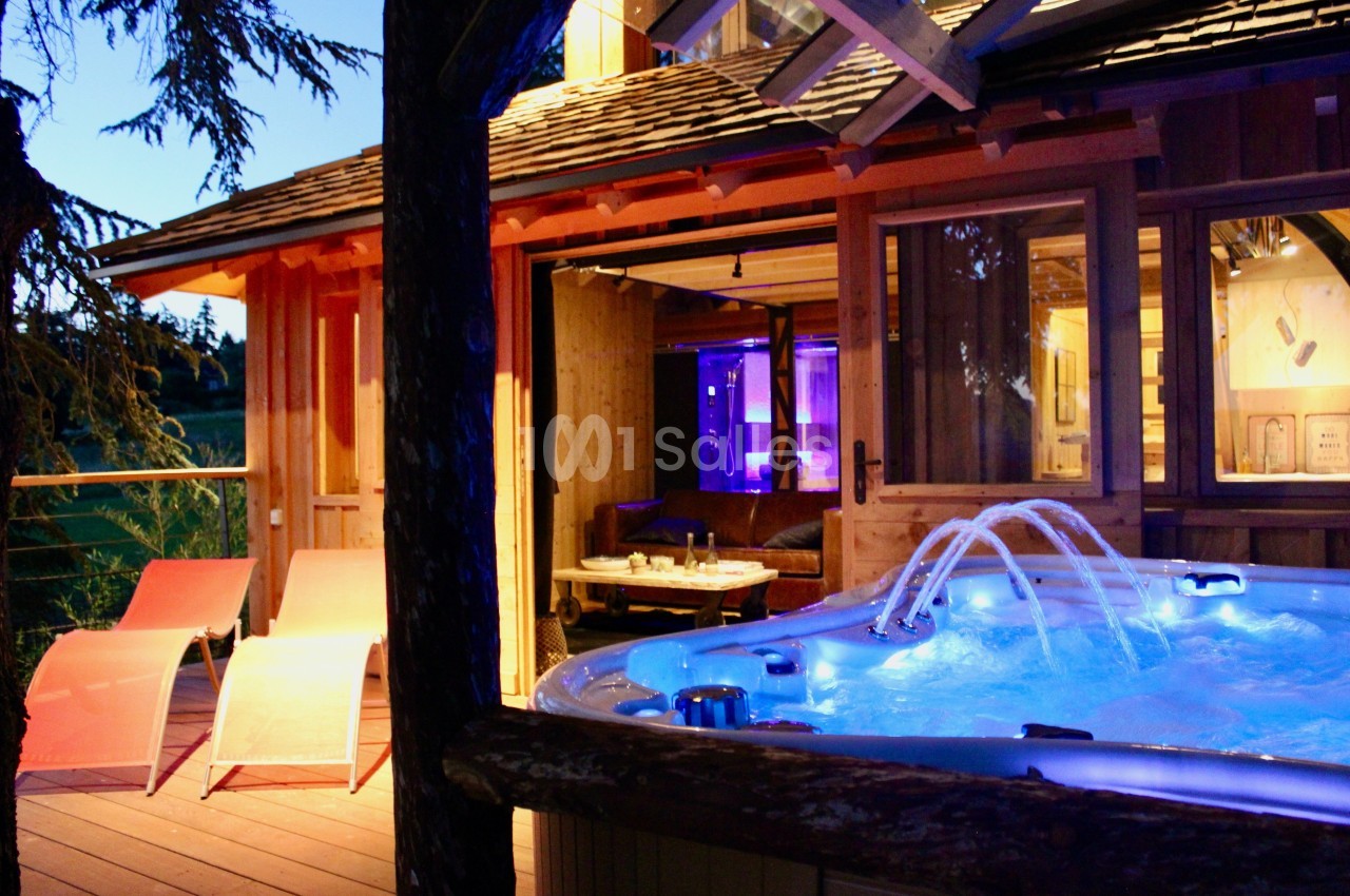 Terrasse en bois avec bain à remous éclairé, transats et vue sur un chalet illuminé en soirée.