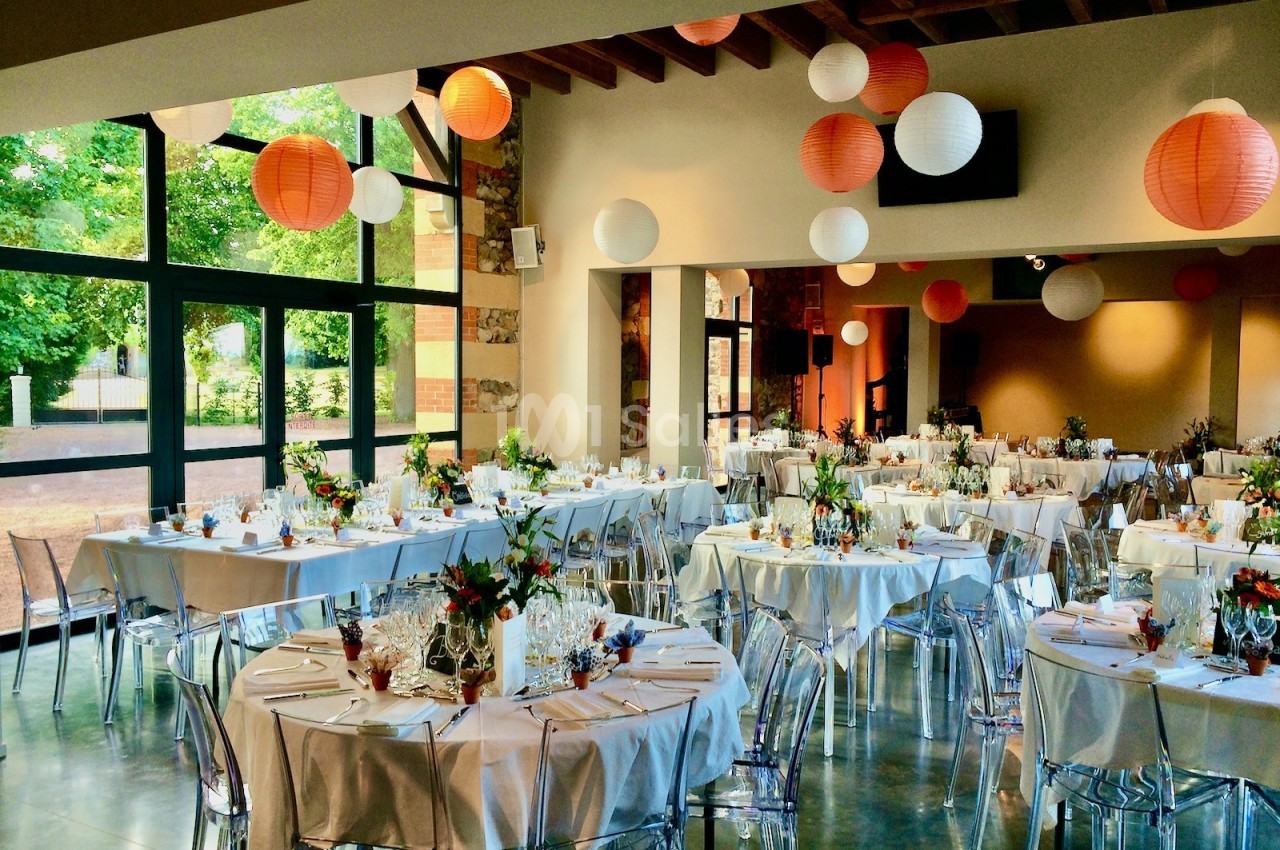 Salle de réception lumineuse avec tables dressées, décorée de lanternes suspendues et de bouquets colorés.