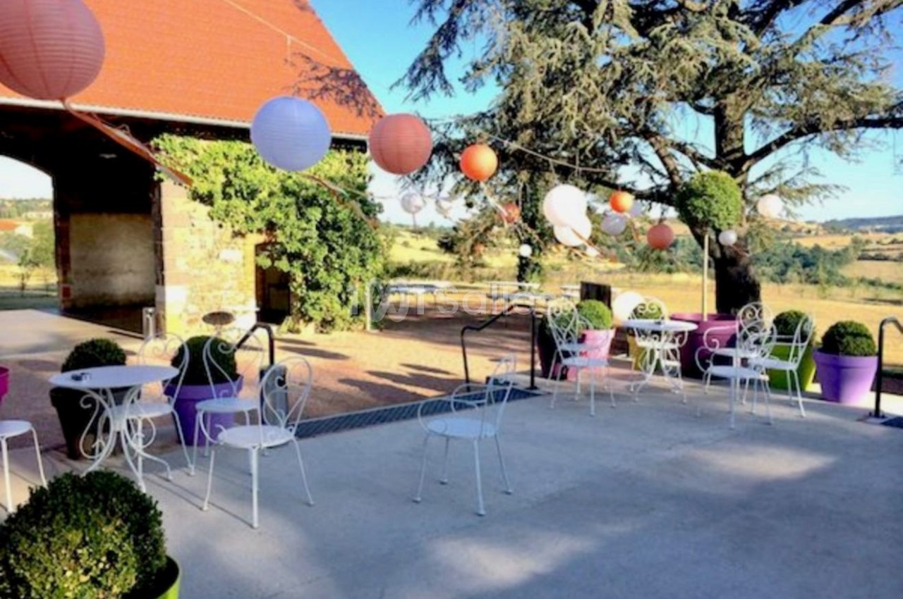 Terrasse extérieure avec tables et chaises en fer forgé, décorée de lanternes suspendues et entourée de verdure.