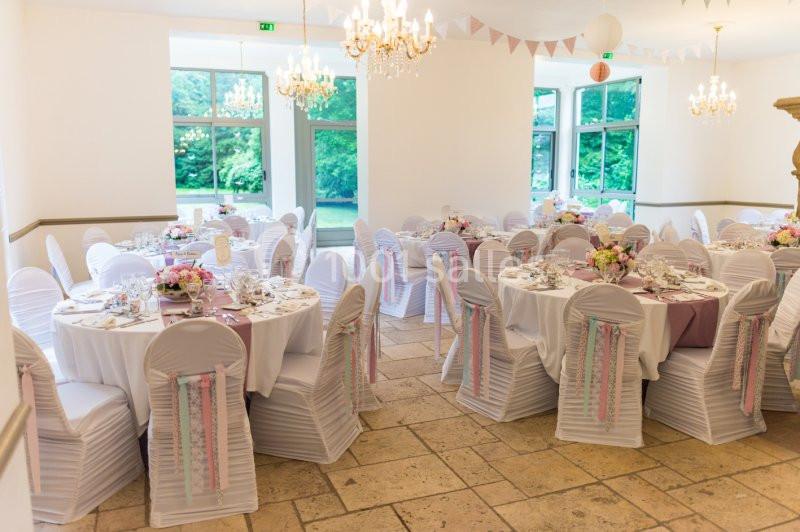 Salle de réception décorée avec des tables rondes, nappes blanches, chaises ornées de rubans et bouquets floraux.
