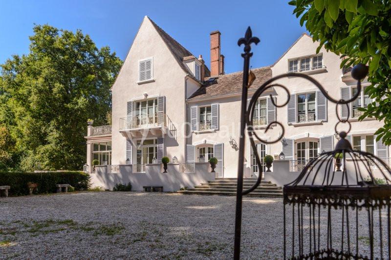 Façade d'une grande maison blanche avec volets, entourée d'arbres, vue depuis une cour gravillonnée.