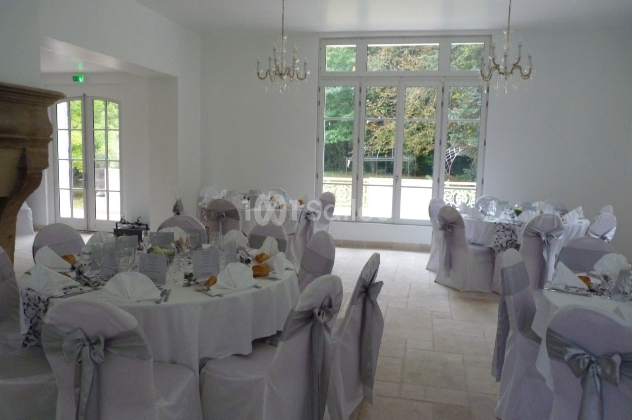 Salle de réception lumineuse avec tables rondes dressées, nappes blanches et chaises couvertes de housses grises.