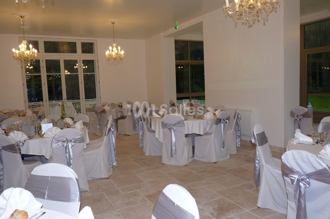 Salle de réception élégante avec tables rondes dressées, chaises décorées de nœuds argentés et lustres suspendus.