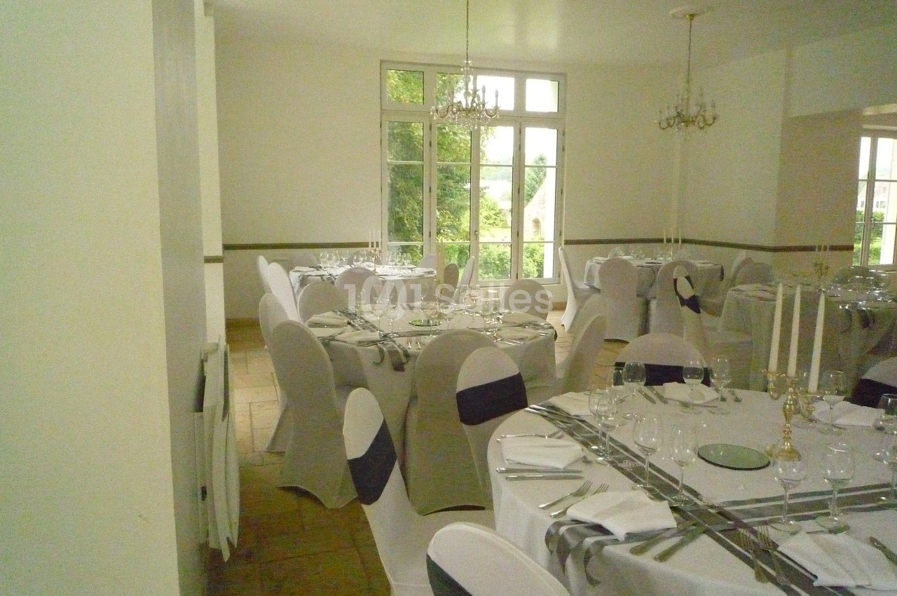 Salle de réception lumineuse avec tables rondes dressées, nappes blanches et chaises couvertes de housses grises.