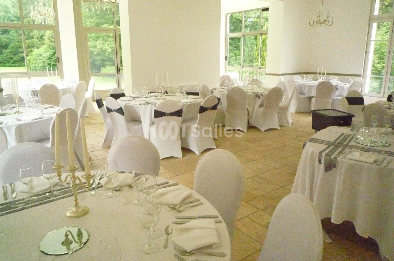 Salle de réception lumineuse avec tables rondes dressées, nappes blanches et chaises couvertes, vue sur un jardin verdoyant.