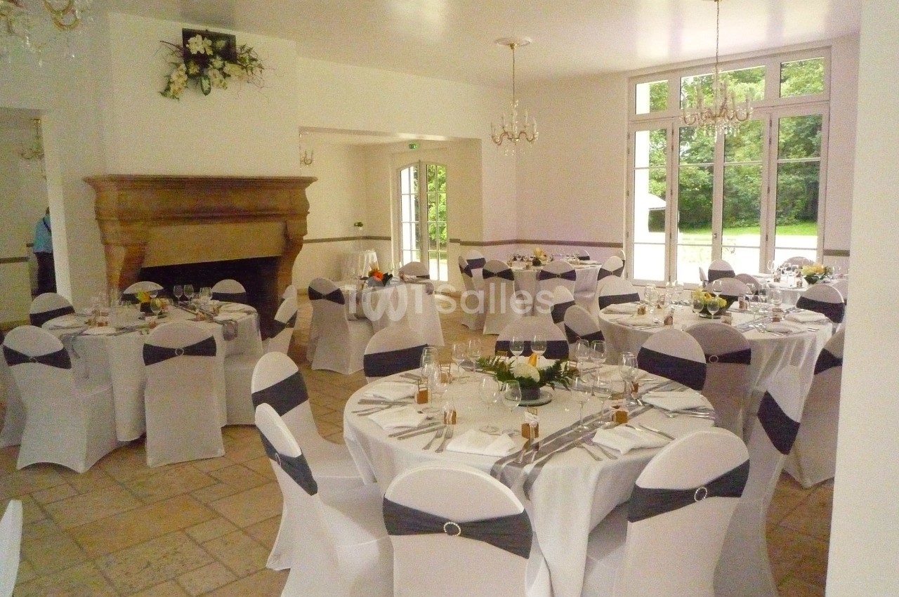 Salle de réception lumineuse avec tables rondes dressées, nappes blanches et chaises décorées de nœuds gris.