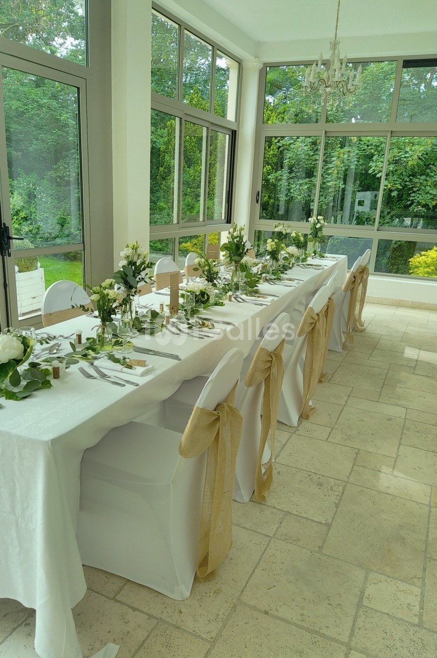 Table de réception décorée avec nappes blanches, chaises ornées de rubans beige et compositions florales, dans une salle…