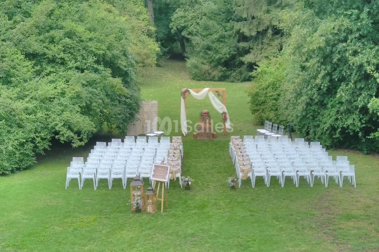 Chaises blanches disposées en extérieur devant une arche décorée de tissus et fleurs, entourées de verdure.