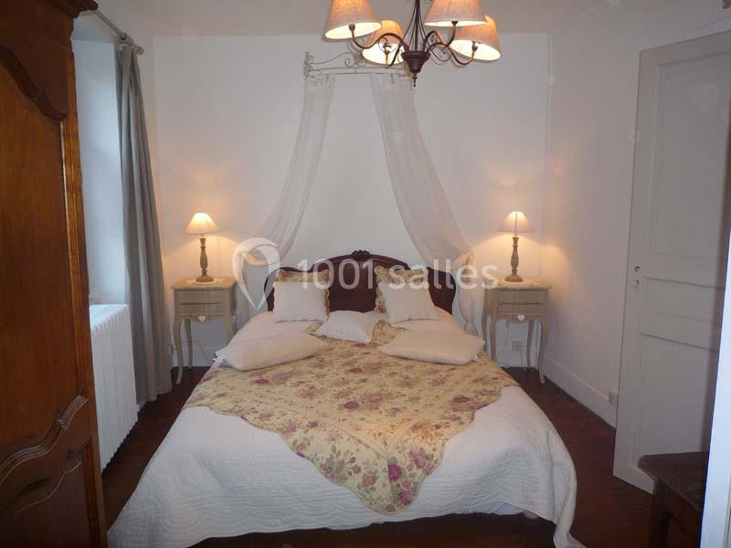 Chambre avec lit double, couvre-lit fleuri, tables de chevet, lampes allumées et rideaux gris, éclairée par un lustre.