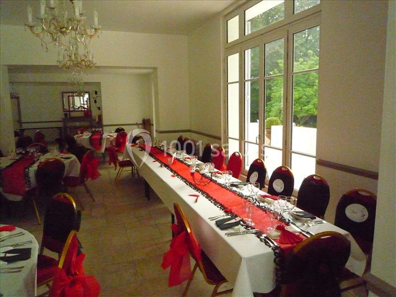 Salle de réception avec de longues tables dressées, nappes blanches et décorations rouges, éclairée par des fenêtres.