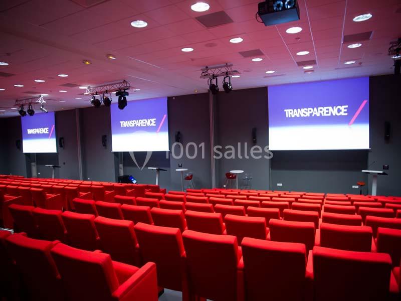 Salle de conférence avec des rangées de sièges rouges et plusieurs écrans affichant le mot ’Transparence’.