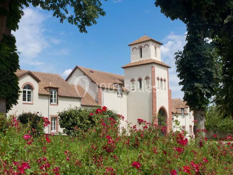 Bâtiment blanc avec une tour à toit en tuiles, entouré de rosiers rouges et d'arbres sous un ciel bleu.