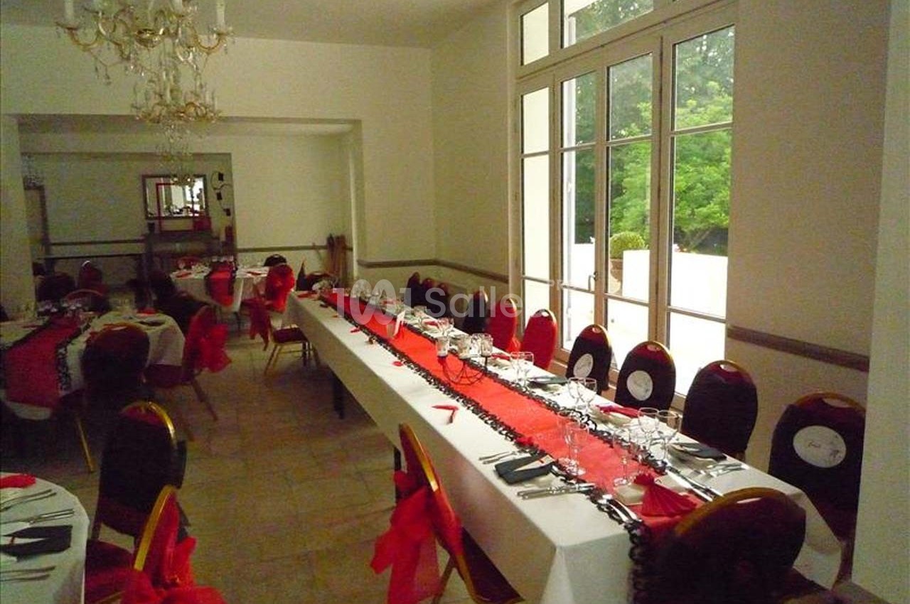 Salle de réception avec tables dressées, nappes blanches et chemins de table rouges, chaises décorées de nœuds rouges.