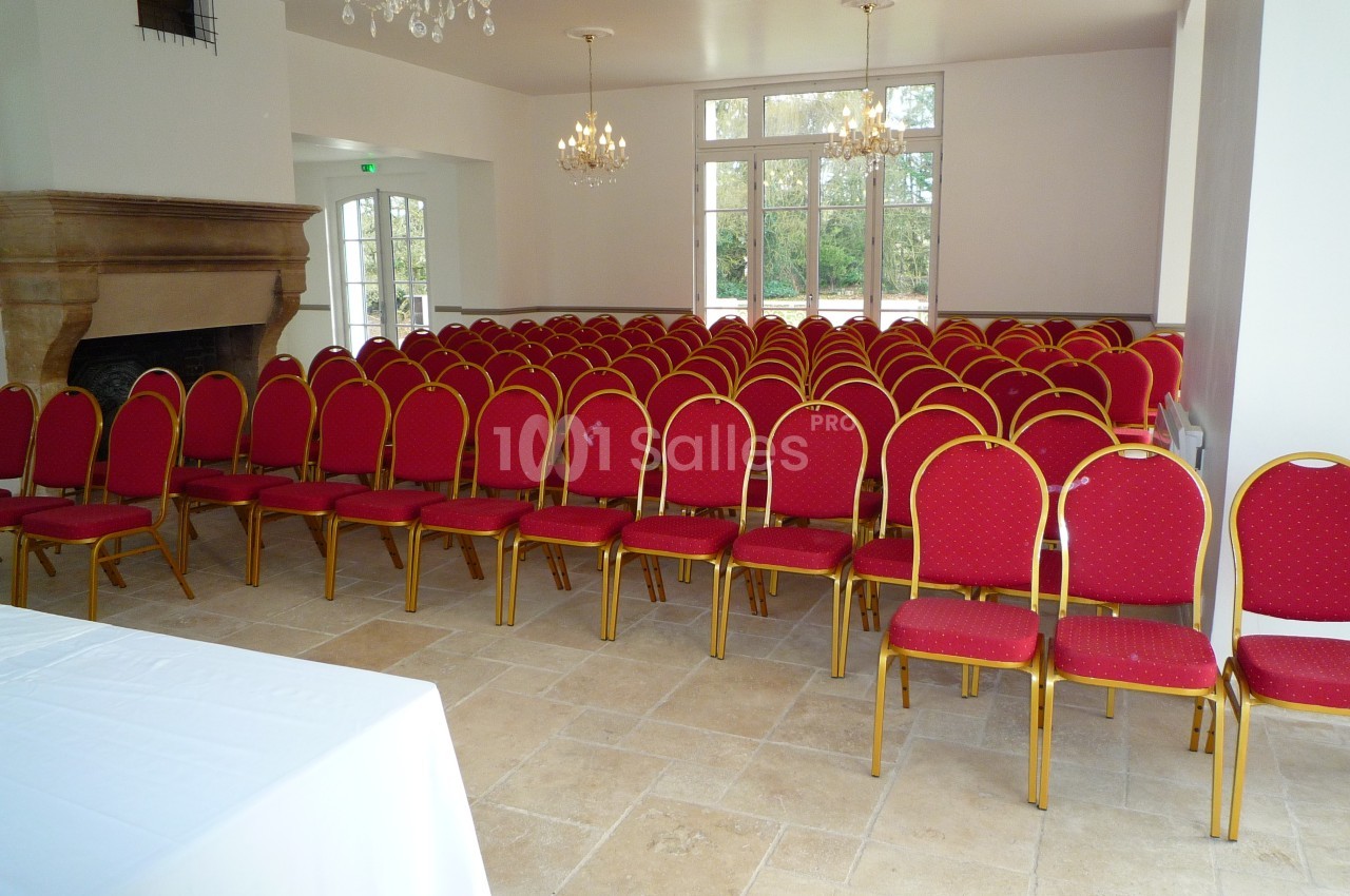 Salle lumineuse avec des rangées de chaises rouges alignées devant une table blanche, cheminée et lustres au plafond.