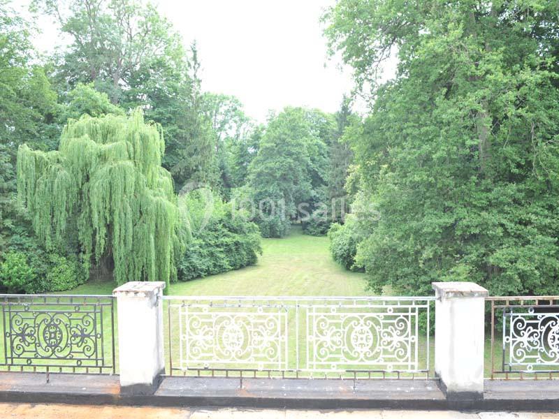 Vue d'un jardin verdoyant avec pelouse, arbres feuillus et un saule pleureur, depuis une terrasse ornée de balustrades en…